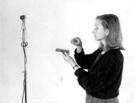 Glödlampa på stativ och en kvinna som håller en plastbit i ena handen och en polaroid i den andra. Foto.